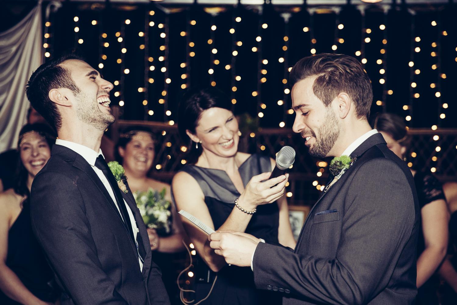 Wedding with two grooms where one is talking into female celebrants microphone, while other groom throws head back in laughter. 