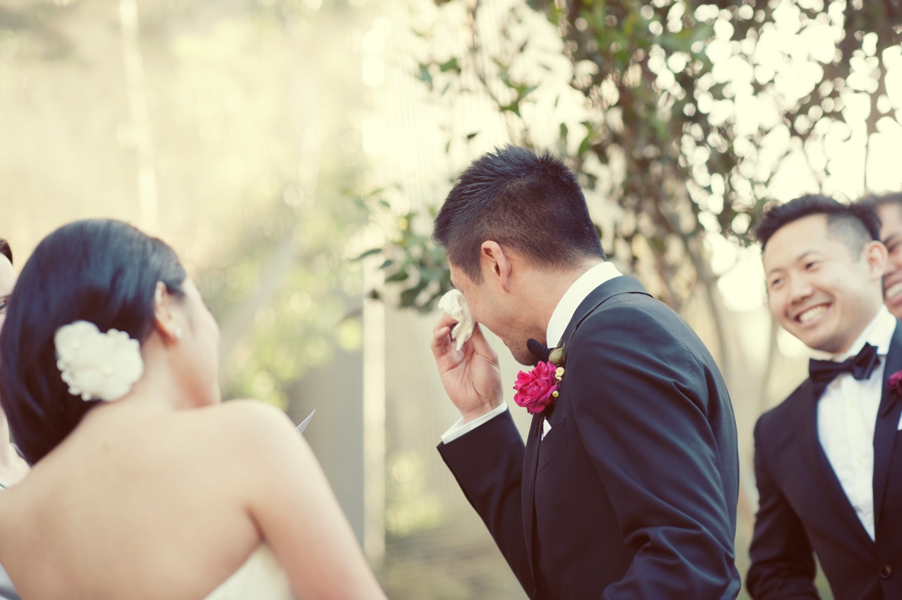 Groom getting emotional during the wedding ceremony and turning away from bride to wipe his eyes with a tissue, while best man watches on and laughs