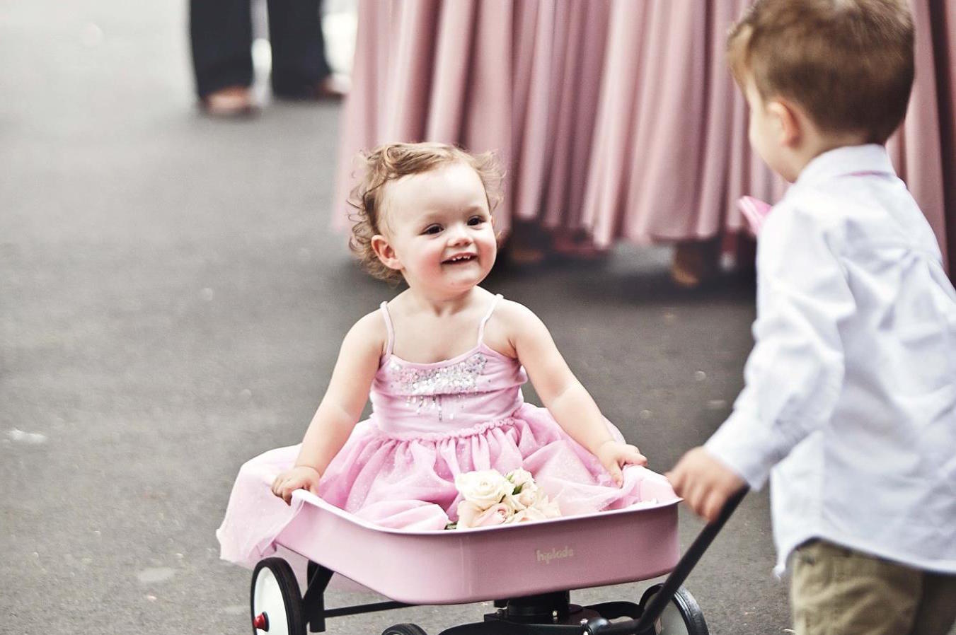 Little smiling flowergirl in pink dress gets pulled down the aisle in a pink wheelbarrow by small boy in shirt and pants