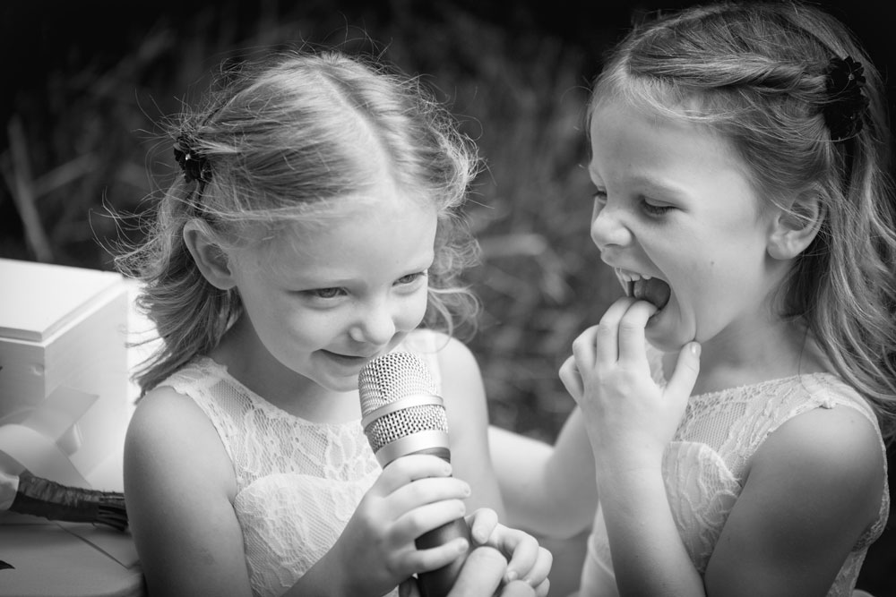 Two young flowergirls laughing and talking into microphone at wedding 