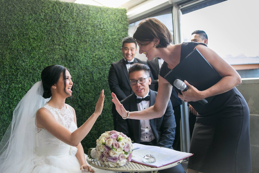 Female celebrant high fives bride during register signing as groom and groomsmen look on 