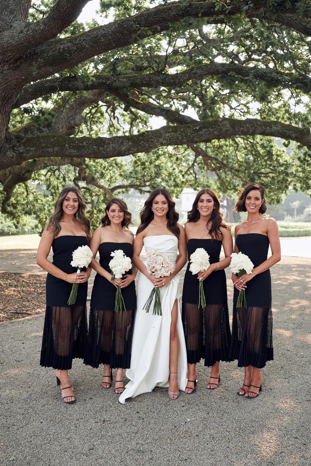 Smiling bride in white and bridesmaids in black, standing in a row under a tree and holding white bouquets.