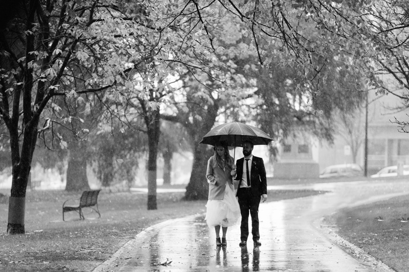 Couple walking alone through a park in the rain with an umbrella on their wedding day 