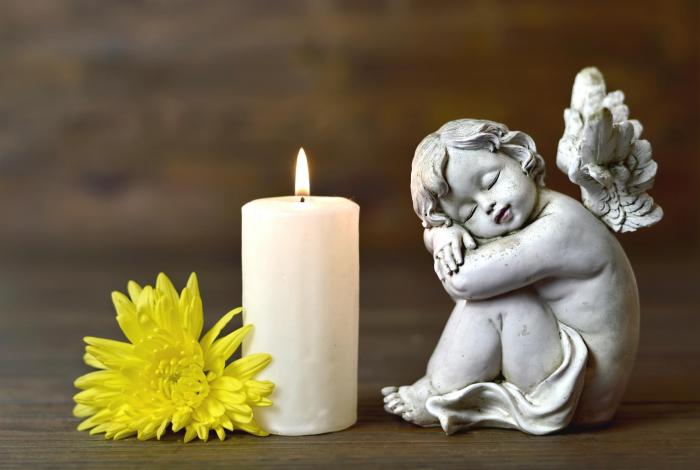 A stone guardian angel positioned next to a single yellow flower and a burning white candle