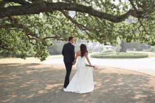 Bride + groom walking away, hand in hand, under a large tree
