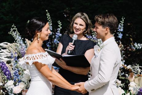 Celebrant holding microphone, smiling at bride and groom during marriage ceremony