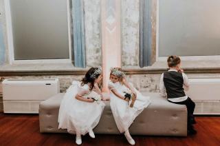 2 flowergirls in white talking together on a cream couch at wedding venue