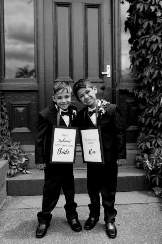 2 young smiling boys dressed in black suits carrying wedding signs around their necks at wedding ceremony