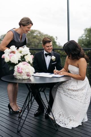 Bride signing the marriage register during wedding ceremony while groom and female celebrant watch on
