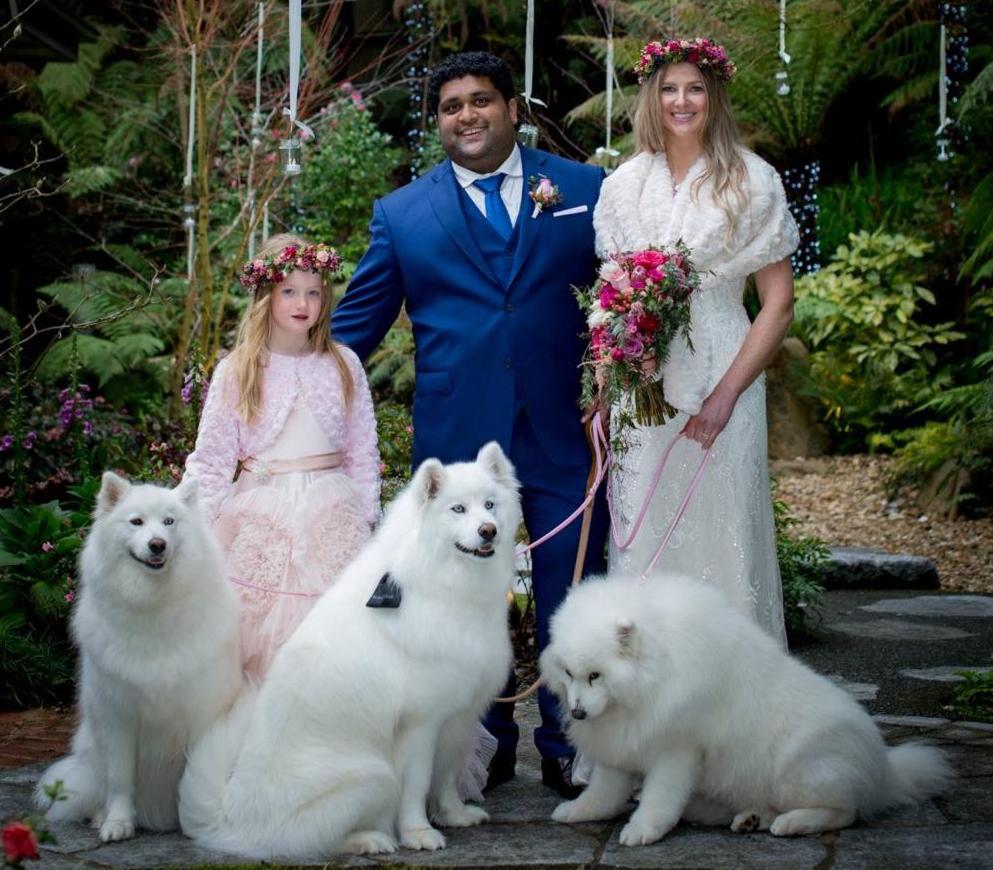 Kids and pets at weddings. Three white fluffy dogs and flowergirl in pink standing with bride and groom
