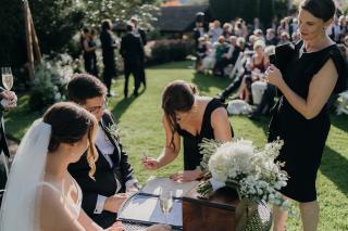 Bride, groom and female celebrant watching witness sign the register at outdoor wedding
