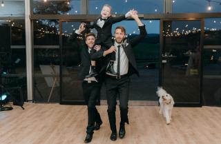 Young page boy gets carried into wedding on the shoulders of two groomsmen with a cream coloured dog running alongside