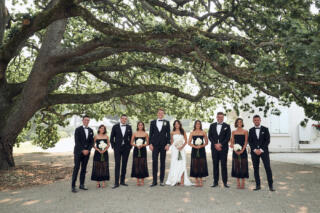 All dressed in black wedding party with bride in white standing in a row under tree