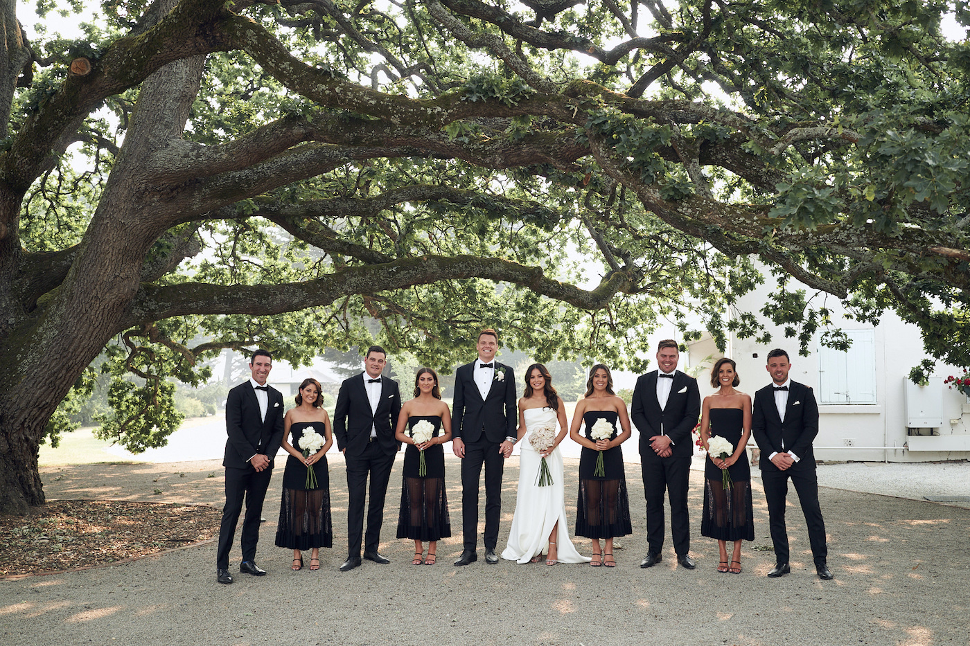 All dressed in black wedding party with bride in white standing in a row under tree 